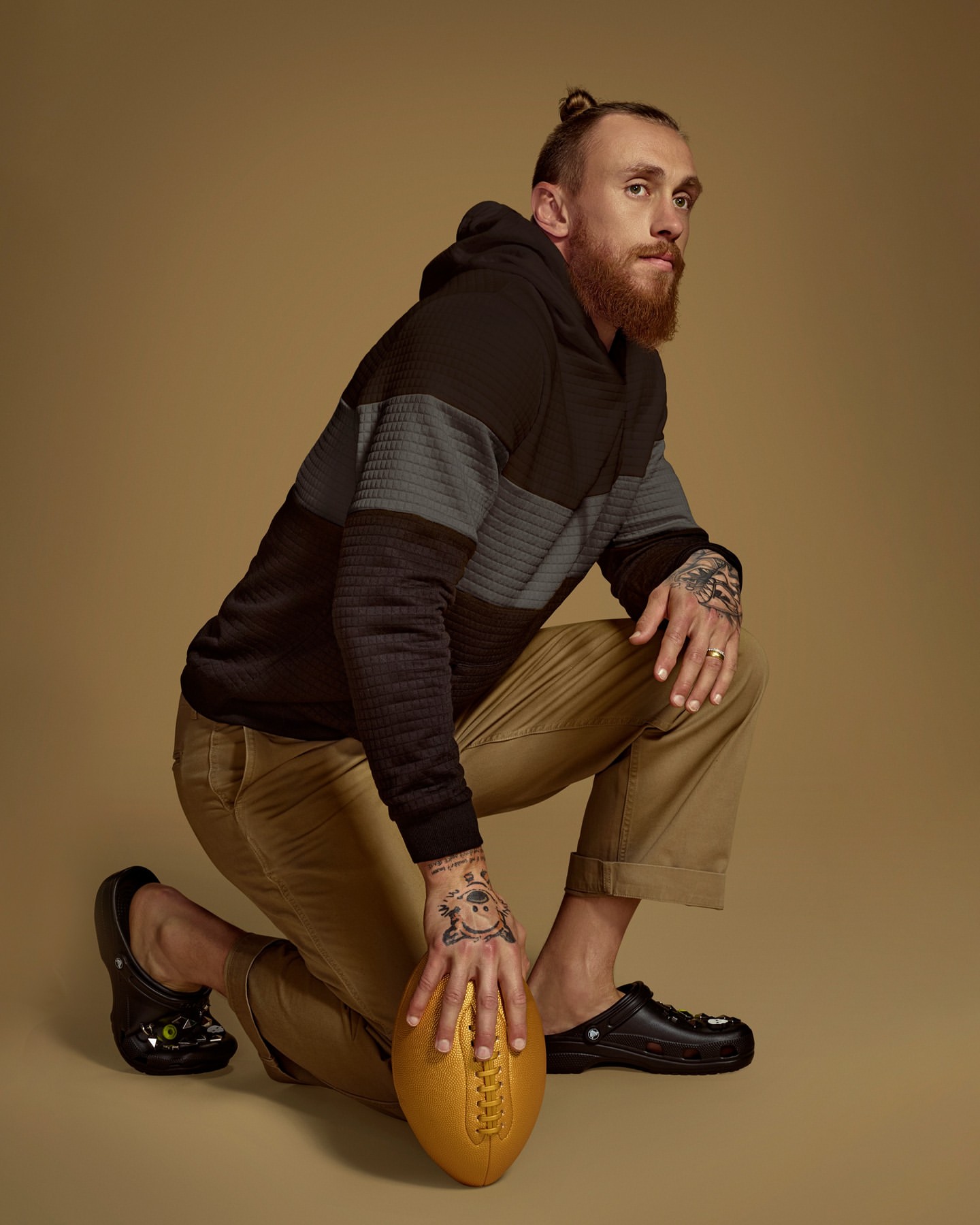 A man with a beard and hair in a bun kneeling in a studio with an american football. He's wearing casual clothes and Crocs footwear.